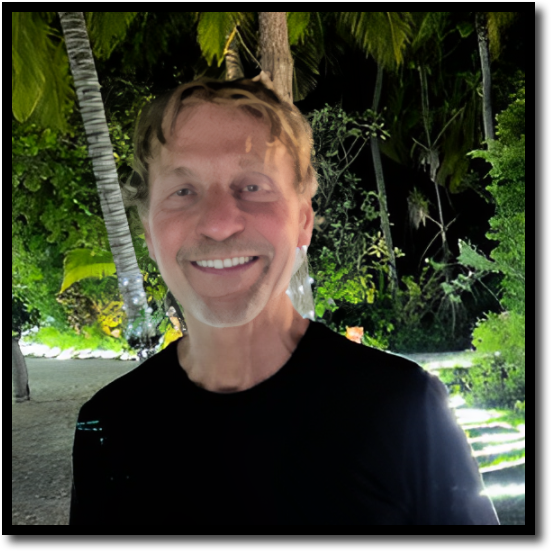 A man smiling outdoors at night, wearing a black shirt, standing on a path surrounded by illuminated palm trees and lush greenery.